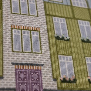 Puede incluir: Bordado de punto de cruz de una fila de casas con una puerta morada, moldura verde y ventanas blancas. Las casas est&aacute;n bordadas sobre una tela beige.