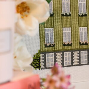 Puede incluir: Un bordado de punto de cruz enmarcado que representa un edificio verde con ventanas blancas y un borde blanco. El edificio tiene un peque&ntilde;o arbusto verde delante. El marco es blanco y el bordado de punto de cruz est&aacute; rodeado por un passe-partout blanco.