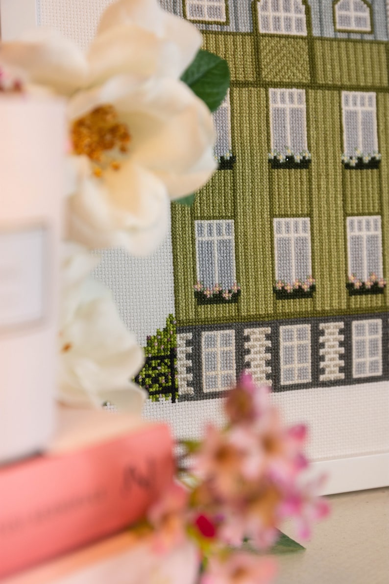 Puede incluir: Un bordado de punto de cruz enmarcado que representa un edificio verde con ventanas blancas y un borde blanco. El edificio tiene un peque&ntilde;o arbusto verde delante. El marco es blanco y el bordado de punto de cruz est&aacute; rodeado por un passe-partout blanco.