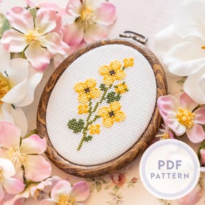 Peut inclure: Broderie au point de croix de fleurs jaunes dans un cercle ovale en bois. Le motif floral est sur tissu blanc. Le cercle est entouré de fleurs roses et blanches. Une étiquette de motif PDF est visible.