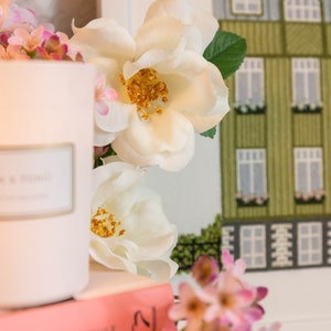 Puede incluir: Una vela blanca con una etiqueta que dice "The Home" se encuentra sobre una pila de libros rosas. Flores blancas con centros amarillos y flores rosas est&aacute;n dispuestas alrededor de la vela y los libros.