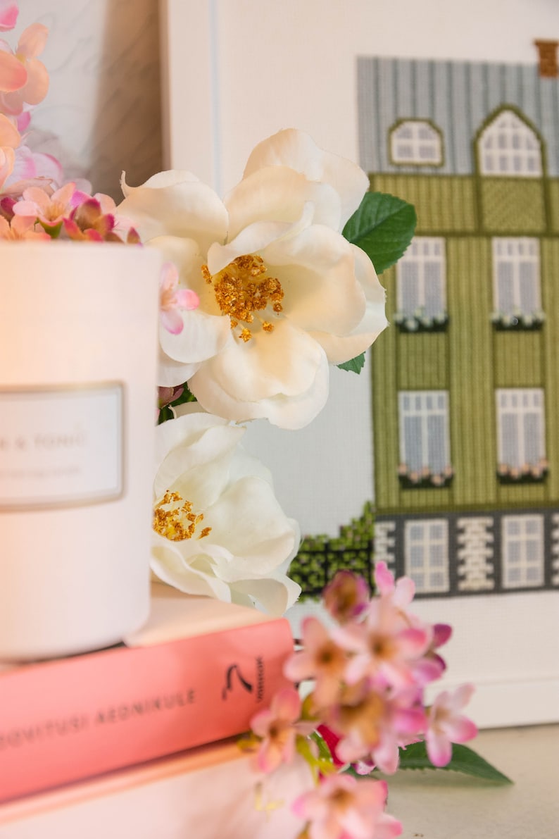 Puede incluir: Una vela blanca con una etiqueta que dice "The Home" se encuentra sobre una pila de libros rosas. Flores blancas con centros amarillos y flores rosas est&aacute;n dispuestas alrededor de la vela y los libros.