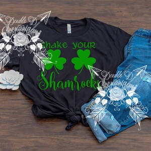 May include: Black t-shirt with the text "Shake your Shamrocks" in green, along with two green shamrock graphics. The shirt is paired with blue jeans on a wooden surface. A decorative floral design is on the left and right.