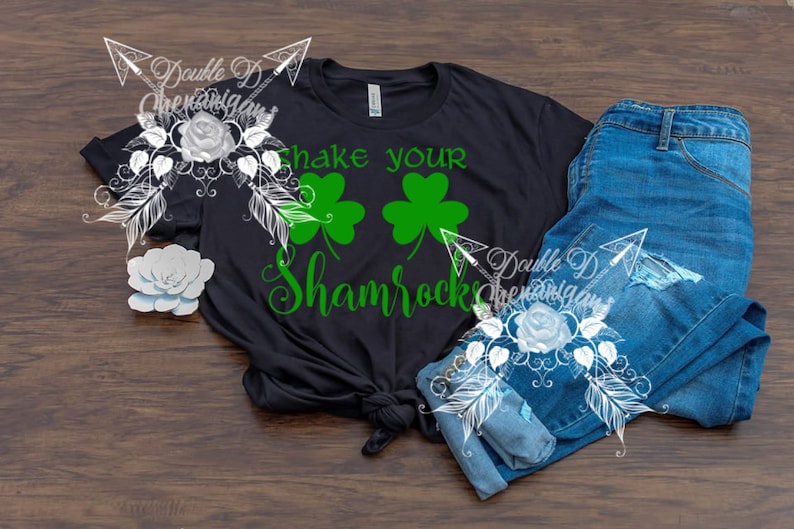 May include: Black t-shirt with the text "Shake your Shamrocks" in green, along with two green shamrock graphics. The shirt is paired with blue jeans on a wooden surface. A decorative floral design is on the left and right.