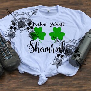 May include: White t-shirt with the text "shake your Shamrocks" and two green shamrock graphics. The shirt has a tied knot at the bottom. A pair of gray and blue hiking shoes and a black water bottle are also in the image.