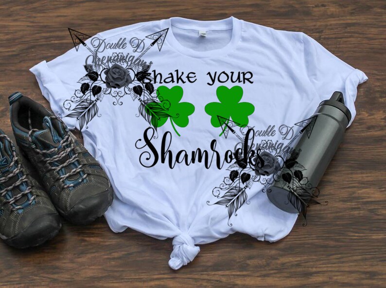May include: White t-shirt with the text "shake your Shamrocks" and two green shamrock graphics. The shirt has a tied knot at the bottom. A pair of gray and blue hiking shoes and a black water bottle are also in the image.