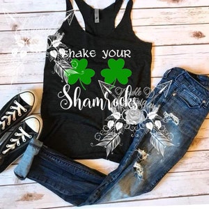 May include: A black tank top with the text "Shake Your Shamrocks" and two green shamrocks. The tank top is paired with distressed blue jeans and black canvas sneakers. The image is set on a light wood background.