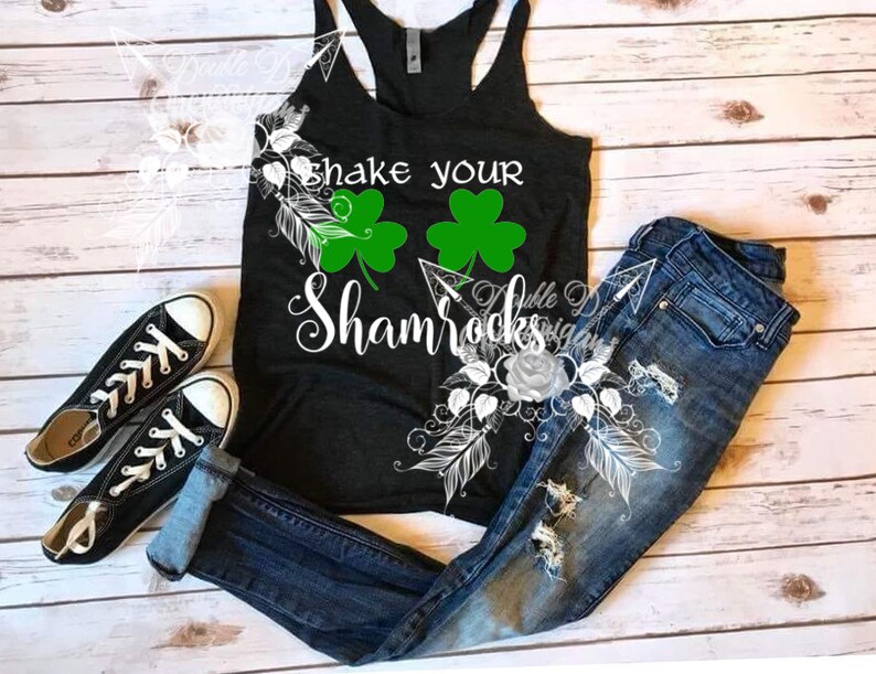 May include: A black tank top with the text "Shake Your Shamrocks" and two green shamrocks. The tank top is paired with distressed blue jeans and black canvas sneakers. The image is set on a light wood background.