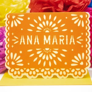 May include: A rectangular orange card featuring intricate laser-cut floral patterns along the edges and the name ANA MARIA centered in bold, cut-out lettering. The card has a scalloped border and is positioned against a backdrop of colorful paper flowers.