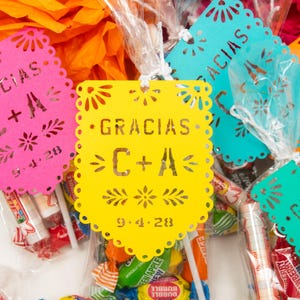 May include: A collection of colorful paper tags in pink, yellow, turquoise, and orange, each featuring die-cut scalloped edges and floral patterns. The text "GRACIAS", "C + A", and "9.4.28" is printed on each tag, which are attached to clear bags of candy.
