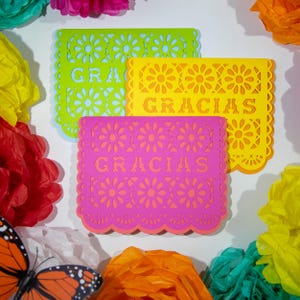 May include: Three colorful paper cutouts with the word "GRACIAS" in orange. The cutouts are green, yellow, and pink, with floral and scalloped edge designs. Surrounding the cutouts are colorful tissue paper flowers and a butterfly.