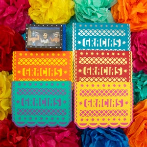 May include: A collection of rectangular paper cards featuring intricate cut-out patterns and the word GRACIAS in bold lettering, presented in various colors including blue, red, orange, and yellow, alongside a small box containing three figurines.