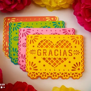 May include: A set of four rectangular paper cards with intricate laser-cut edges and a central heart design. Each card features the word GRACIAS in the center. The cards are layered in yellow, pink, green, and orange, surrounded by colorful paper flowers.