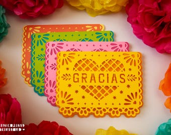 May include: A set of four rectangular paper cards with intricate laser-cut edges and a central heart design. Each card features the word GRACIAS in the center. The cards are layered in yellow, pink, green, and orange, surrounded by colorful paper flowers.