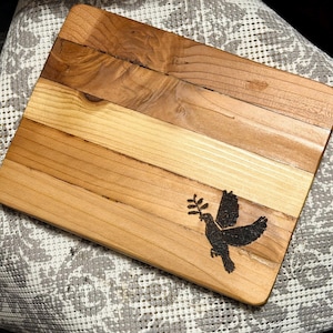 May include: Wooden chopping board featuring a dove design. The rectangular board is made of light and dark wood planks. A black dove carrying an olive branch is burned into the wood. The board is approximately 30 cm long and 20 cm wide.