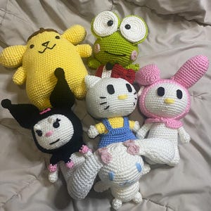 May include: A collection of six handmade crochet plush toys. The toys include a yellow dog-like character, a green frog, a white cat with a red bow, a pink bunny, a black character with a pink bow, and a white character with blue accents.