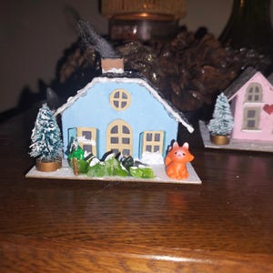 Puede incluir: Una casa azul en miniatura con techo blanco, nieve y una pequeña figura de zorro naranja. La casa tiene una ventana redonda y una chimenea. Un pequeño árbol de Navidad está a la izquierda. Tema de pueblo de invierno.