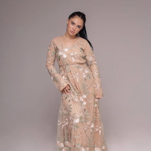 Puede incluir: Una mujer con un vestido largo de color beige con bordados florales. El vestido tiene un escote en V y mangas largas. La mujer está de pie sobre un fondo blanco.