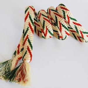 May include: A white, green and red braided cord with tassels at each end. The cord is woven in a chevron pattern.
