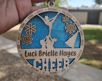 Op de afbeelding: Een houten ornament met een blauwe glitterachtergrond en een silhouet van een cheerleadingteam. De ornament is versierd met sneeuwvlokken en de woorden "Luci Brielle Hayes" en "CHEER". De ornament bevat ook een lijst met positieve eigenschappen die geassocieerd worden met cheerleading, zoals "atletisch", "gepassioneerd", "teamgericht", "geconcentreerd", "taai", "geestig", "vastberaden", "competitief", "hart", "gecommitteerd", "fit" en "consistent".