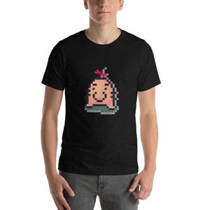 May include: A black t-shirt with a pixelated image of a pink creature with a red flower on its head. The creature has a smiling face and is wearing a green hat.