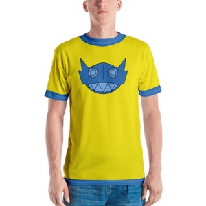 Può includere: Una maglietta gialla con un design geometrico blu e bianco sul davanti. Il design presenta un viso stilizzato con due occhi grandi e una bocca larga.