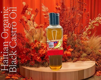 May include: A bottle of Haitian Organic Black Castro Oil with a red, white, and blue label. The label features a banana and the text "Black Castro Oil". The bottle is sitting on a wooden surface in front of a red curtain.