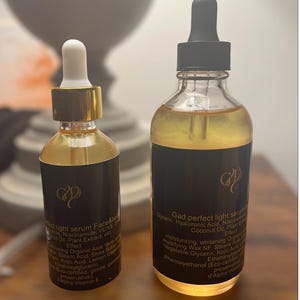May include: Two glass bottles of serum with black labels and gold accents. The bottles contain a golden liquid and have droppers. The labels include text about the serum's ingredients and effects. The bottles are on a light-colored surface.