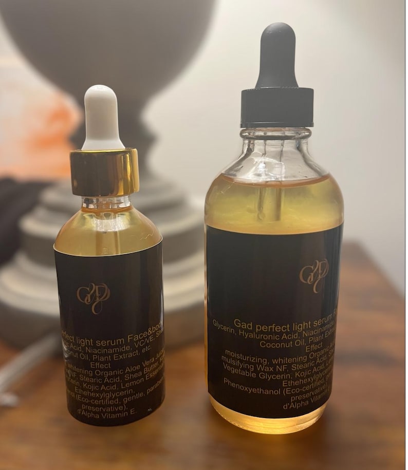 May include: Two glass bottles of serum with black labels and gold accents. The bottles contain a golden liquid and have droppers. The labels include text about the serum's ingredients and effects. The bottles are on a light-colored surface.