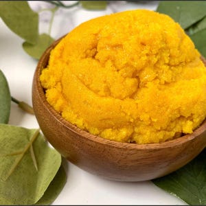 Puede incluir: Primer plano de un exfoliante amarillo, granulado y texturizado en un cuenco de madera. El exfoliante está hecho con ingredientes naturales y es perfecto para exfoliar la piel.
