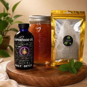 May include: A blue glass bottle labelled Superfood Oil Daily Detox with a chakra graphic, a clear glass mason jar filled with amber liquid, and a gold foil pouch containing a tea bag labelled Miracle Tea, accompanied by a fresh mint sprig.