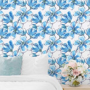 May include: A bedroom scene featuring wallpaper with a repeating pattern of watercolor-style blue flowers on a white background. A light blue pillow and a white duvet are visible in the foreground, with a bouquet of pink and white flowers in a vase.