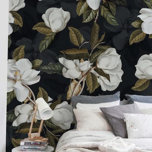 Può includere: Un murale con grandi fiori di magnolia bianchi e foglie verde scuro su sfondo nero. Una lampada da scrivania bianca è appoggiata su una pila di libri accanto a un letto con cuscini grigi e bianchi e una coperta bianca in primo piano.