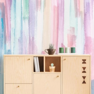 May include: A light-coloured wooden sideboard with two cabinets, a drawer, and open shelving. The cabinet features small round gold knobs and a vertical row of dark wood geometric cutouts. A small plant and three green candles sit on top.