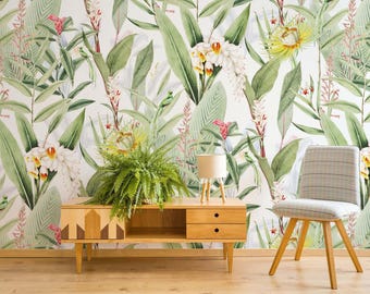 May include: A light wood media console with two drawers and an open shelf sits against a wall covered in botanical wallpaper featuring green leaves, white orchids, and pink flowers. A green fern in a pot and a small lamp sit on the console.
