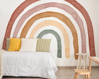 May include: A watercolor rainbow mural in shades of brown, pink, yellow, and green adorns a white wall. Below, a bed with white bedding and decorative pillows in yellow and green sits in front of the mural. A small wooden stool and table are in the foreground.