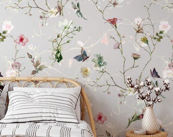 Könnte beinhalten: Ein Schlafzimmer mit hellgrauer Blumentapete mit Schmetterlingen und Blumen in Rosa, Lila und Grün. Im Vordergrund ein Bett mit gestreiftem Kissen und Decke, dazu eine Vase mit Baumwollstielen auf einem kleinen Weidenkorbtisch.