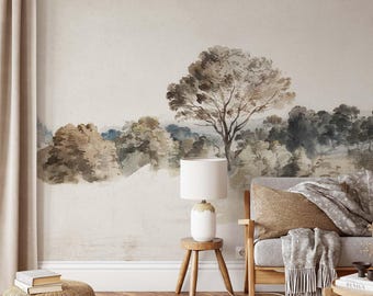 Op de afbeelding: Een muurschildering met een aquarel landschapsschilderij van bomen in tinten bruin, grijs en gedempt blauw tegen een lichtbeige achtergrond. De scène bevat een houten bijzettafel met een witte lamp, een bank met een gestructureerd kussen en een rieten poef.