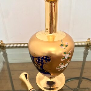 May include: A gold and blue glass decanter with a floral design and a gold stopper. The decanter has a round body and a tall neck.