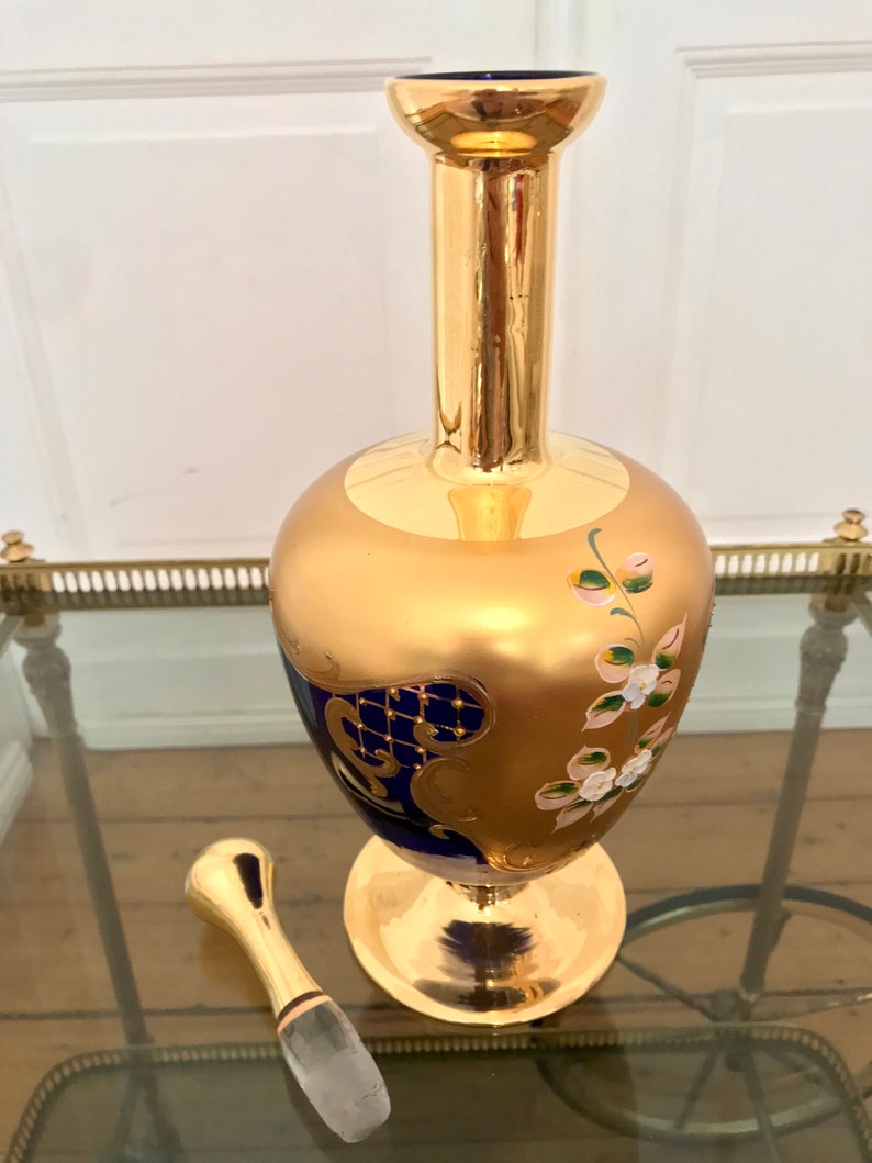 May include: A gold and blue glass decanter with a floral design and a gold stopper. The decanter has a round body and a tall neck.
