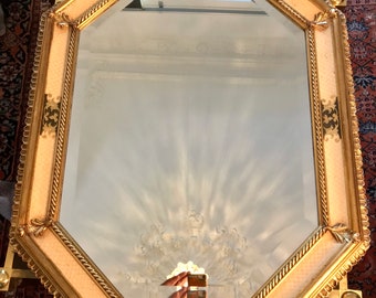 May include: An octagonal mirror with a gold frame and decorative details. The frame features a cream-colored central panel with a patterned design. The mirror is supported by a gold stand, set against a patterned rug.