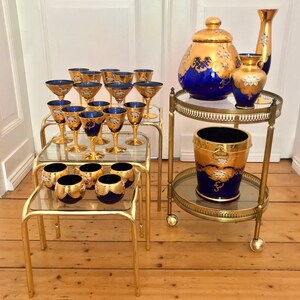 May include: Set of gold and blue glassware with gold accents. The set includes a large pitcher, a smaller vase, and a set of glasses. The glassware is displayed on a gold metal serving cart with glass shelves.