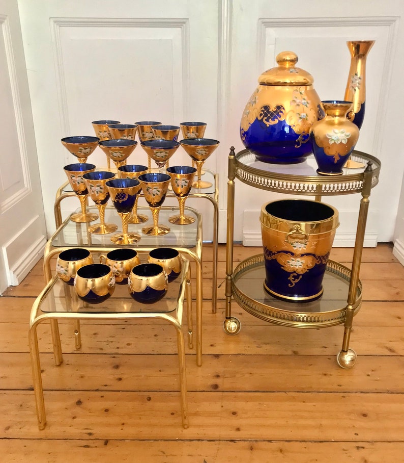 May include: Set of gold and blue glassware with gold accents. The set includes a large pitcher, a smaller vase, and a set of glasses. The glassware is displayed on a gold metal serving cart with glass shelves.