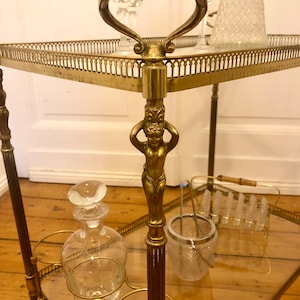 May include: A gold-toned metal serving trolley with glass shelves. The trolley has a decorative handle and a sculpted figure on the front post. The trolley is on a wooden floor.