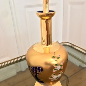 May include: A gold and blue glass decanter with floral and geometric designs. The decanter has a round body and a tall, slender neck.