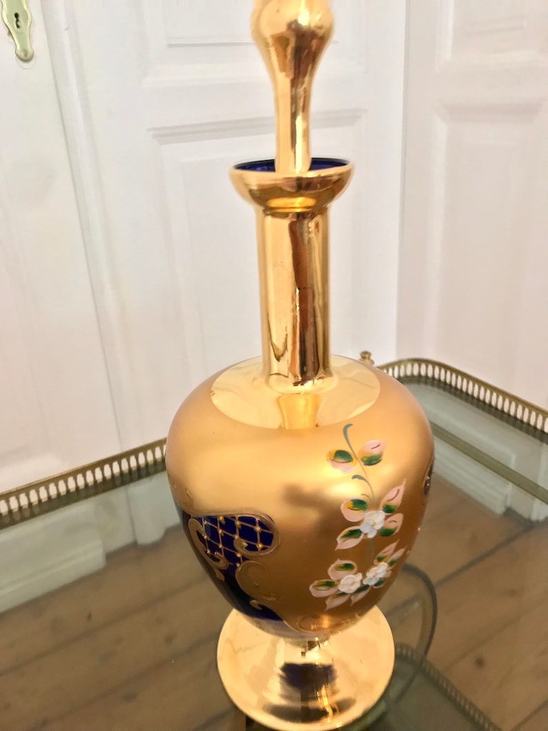 May include: A gold and blue glass decanter with floral and geometric designs. The decanter has a round body and a tall, slender neck.