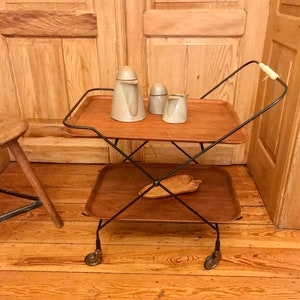 May include: A black metal serving cart with two wooden trays. The cart has wheels and a handle. There are three ceramic containers on the top tray.