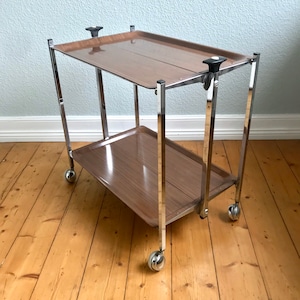 Könnte beinhalten: Ein Servierwagen aus Chrom und Holz mit zwei Ablagen und vier Rädern. Der Wagen hat eine braune Holzplatte oben und eine untere Ablage.