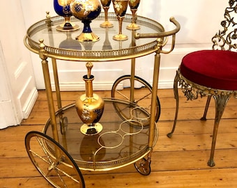 May include: A gold bar cart with glass shelves and wheels. The cart is decorated with gold accents and has a glass top. There are four blue and gold glass decanters and six matching glasses on the top shelf. The cart is on a wooden floor.