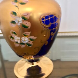 May include: A cobalt blue glass vase with gold accents and floral designs. The vase has a wide base and a narrow neck.